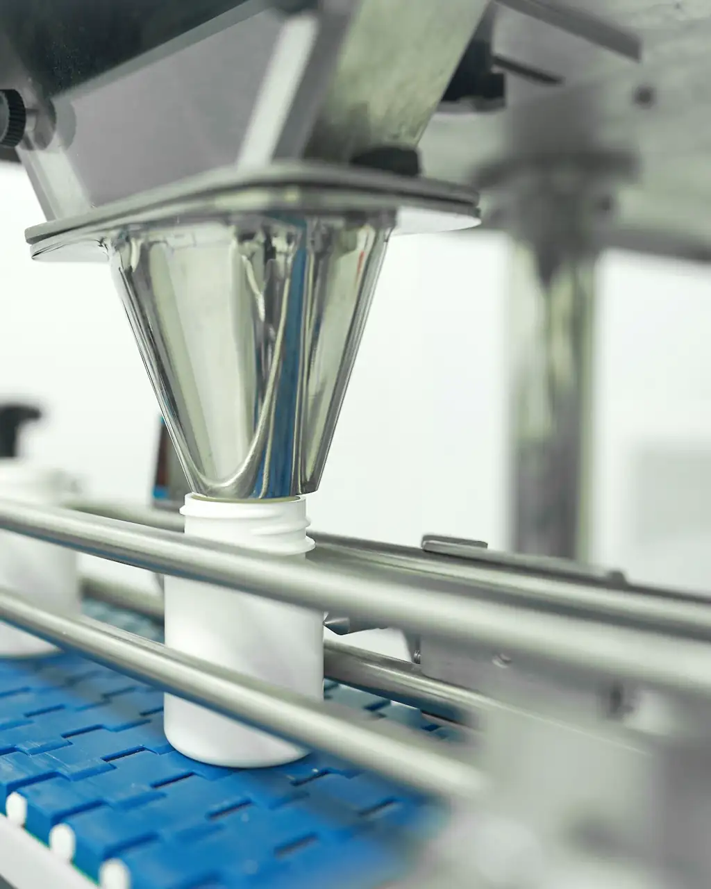 A container moving along a blue conveyor beneath a stainless-steel filling machine on a production line.